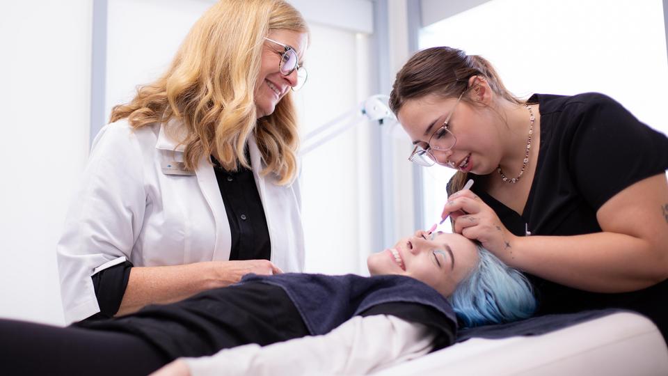 A person laying down while a woman treats their face and another woman looks on.