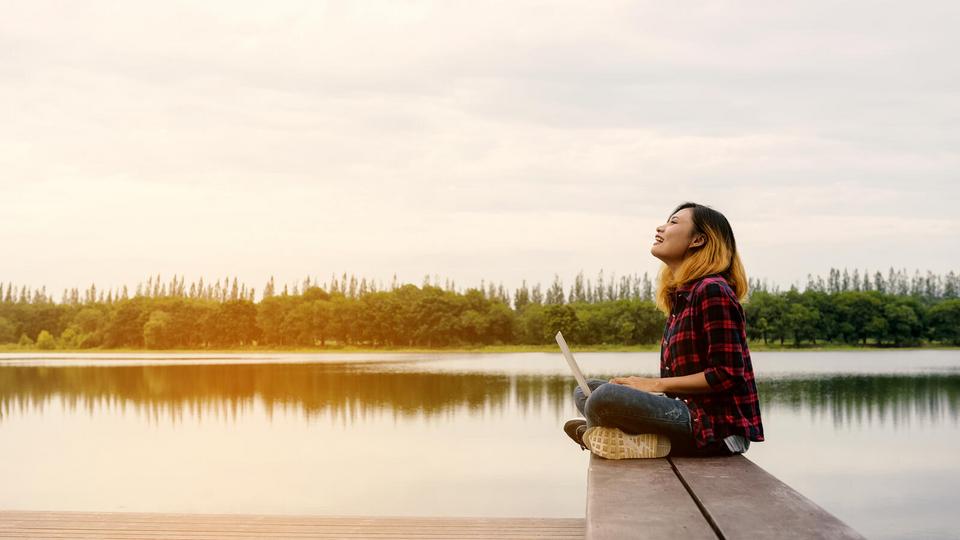 Woman sitting cross-legged with her laptop on her lap on the end of a wooden dock which sits on a reflective lake with a tree line in the background