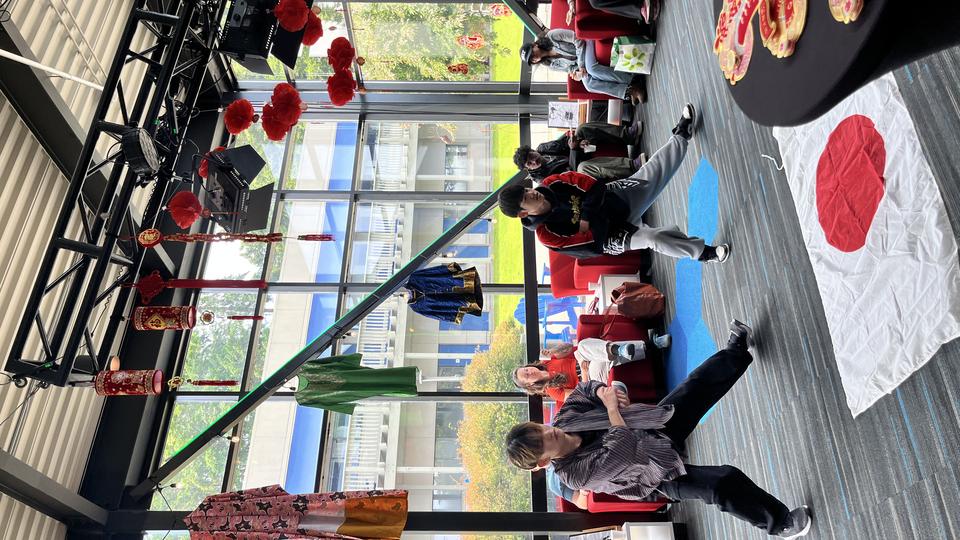 Students perform a dance inside a bright campus lounge decorated for an Asian cultural event, with red lanterns and traditional garments hanging overhead.