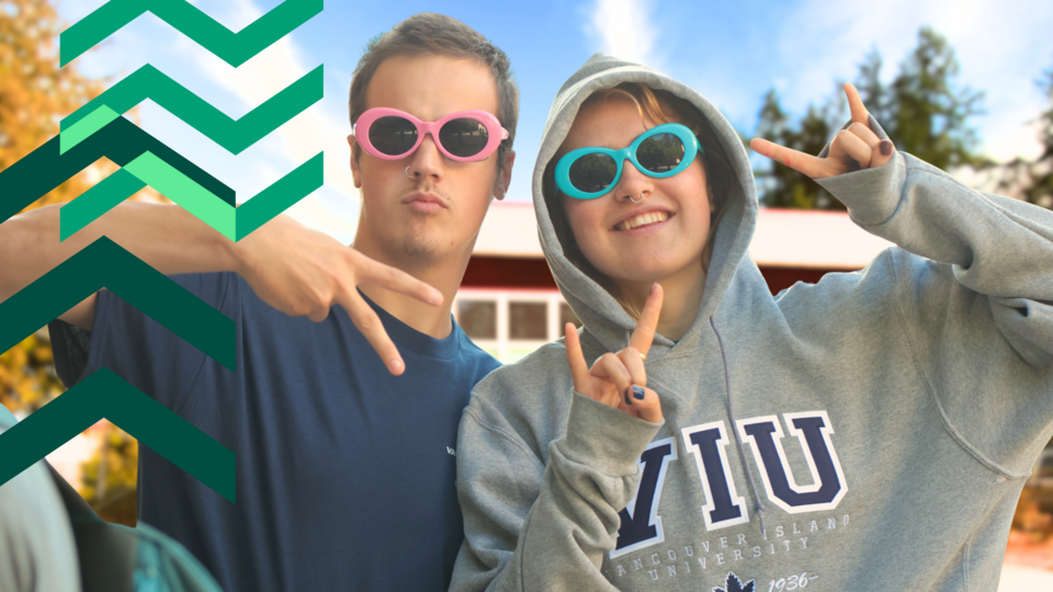 Two students wearing sunglasses pose in front of VIU tiwsemawtxw campus
