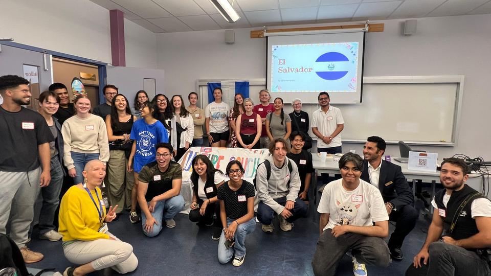 A diverse group of students and staff pose together in a classroom during a Culture Couch event. A screen at the front displays “El Salvador” with the national flag.  Participants are smiling and standing or kneeling in a semi-circle, creating a welcoming and celebratory atmosphere.